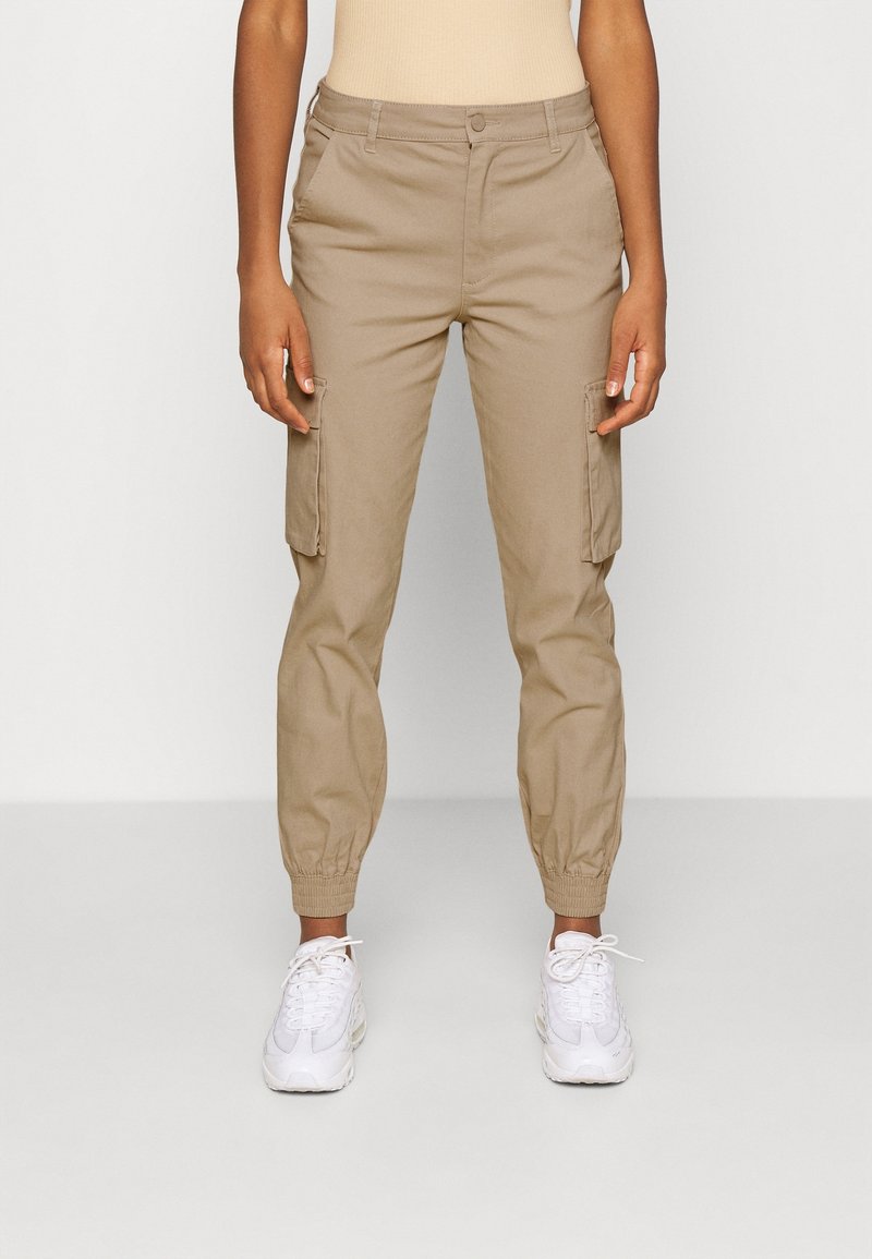 Pantalons cargo beiges avec des poignets élastiques, dotés de poches latérales et d'une texture lisse. Associés à des baskets blanches.