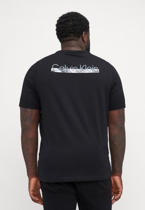 Mężczyzna z tatuażami w czarnej koszulce Calvin Klein, z widocznym logo marki na górnej części pleców, na jednolitym jasnym tle.