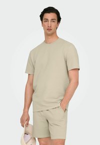 Beige tekstureret strik t-shirt med korte ærmer, parret med matchende shorts, som har en afslappet pasform. Modellen holder en lysfarvet kasket.
