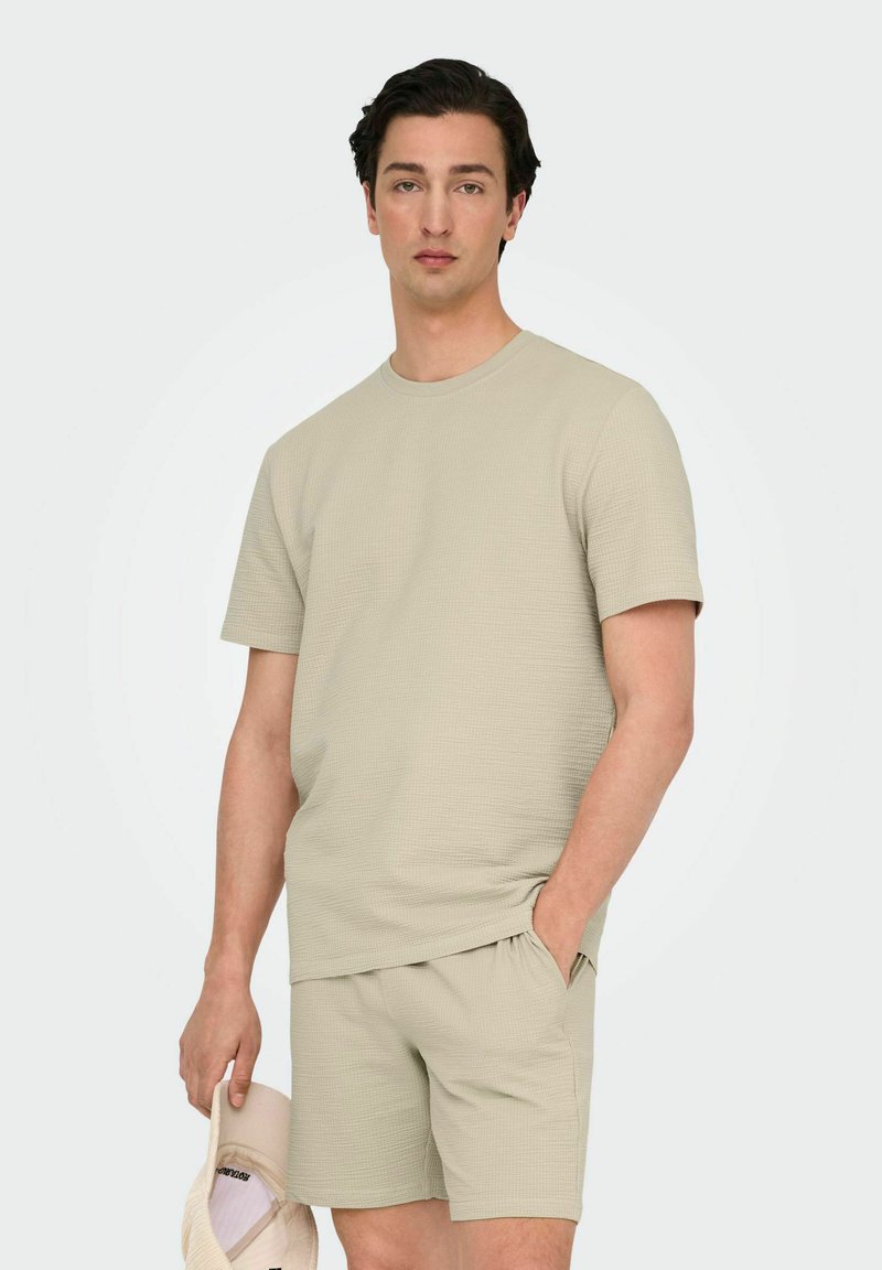 Beige tekstureret strik t-shirt med korte ærmer, parret med matchende shorts, som har en afslappet pasform. Modellen holder en lysfarvet kasket.