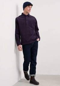 Tumši purpura krāsas kordūrāna krekls ar pogājamu apkakli un priekšējo kabatu, apvienots ar tumšām džinsām un brūnām potīšu zābakiem, uz baltas sienas.