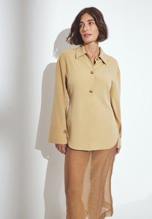 Camisa de manga larga beige con cuello y botones dorados, usada sobre una falda marrón transparente, contra un fondo blanco. Textura de tela suave.