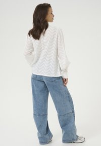 Witte top met zigzagpatroon en lange mouwen, gecombineerd met lichtblauwe denim jeans met een losse pasvorm. De textuur lijkt zacht te zijn, met een ontspannen silhouet.