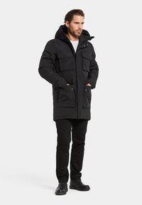 Parka noire rembourrée avec une capuche, équipée de quatre poches avant, d'une fermeture éclair et de boutons-pression. Portée avec un pantalon noir et des bottes.