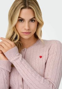 Svijetlo ružičasti kardigan od pletenine sa malim crvenim srcem, rebrastim manžetama i kopčanjem na gumb. Mekana tekstura i prilagođen oblik.