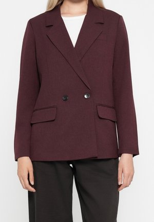 Vrouw die een donker pruimkleurig dubbelreeks blazer draagt met twee klepzakken, over een witte top en zwarte broek, staand tegen een effen achtergrond.