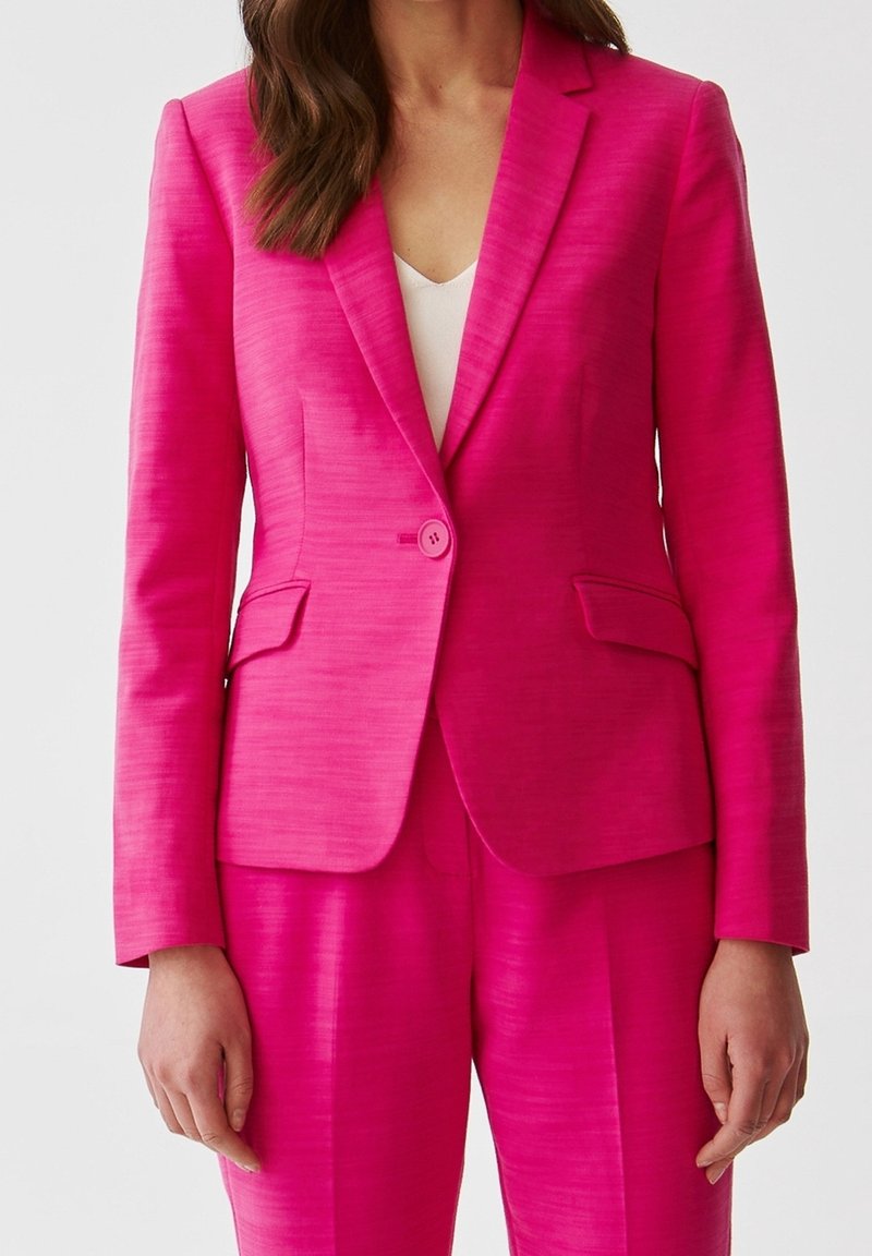 Blazer rose ajusté en tissu texturé, fermeture à un bouton, revers crantés et deux poches latérales. Associé à un pantalon assorti.