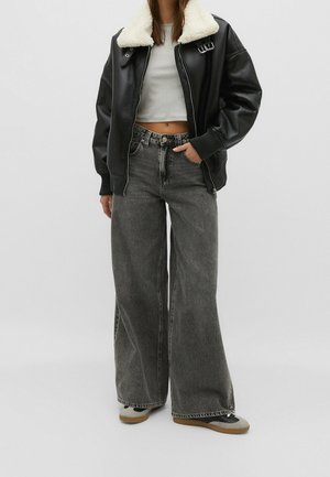 Henkilö, jolla on musta nahkatakki valkoisella karvahihalla, lyhyt valkoinen paita, leveälahkeiset harmaat farkut ja lenkkarit, seisoo käsi taskussa.