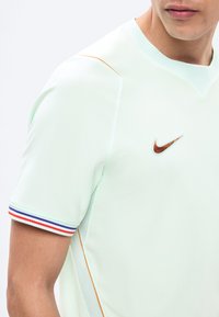 Άνδρας που φοράει ανοιχτό πράσινο αθλητικό μπλουζάκι Nike με πορτοκαλί ρίγα και ριγέ κόκκινο, λευκό και μπλε περιθώριο στο μανίκι.