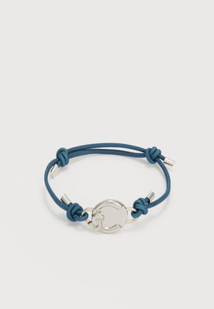 Braccialetto con cordino blu, chiusura circolare in argento e terminali in metallo, fissato con più nodi su uno sfondo neutro.