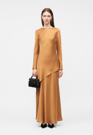 Frau in einem langärmeligen, asymmetrischen, beigefarbenen Kleid, die eine kleine schwarze, mit Fransen verzierte Handtasche hält, schwarze spitze Schuhe trägt und vor weißem Hintergrund steht.