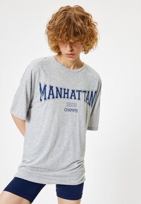 Szara, oversize koszulka z niebieskim napisem "MANHATTAN 1995 CHAMPS". Miękki materiał z bawełny, krótkie rękawy, luźny krój. Noszona z granatowymi szortami.