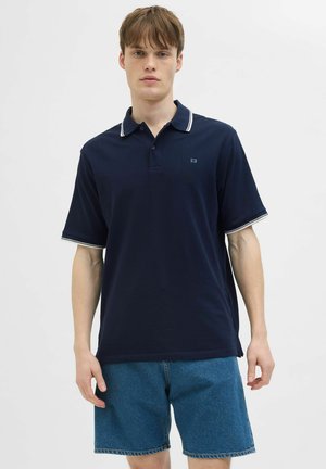 Joven vestido con camiseta tipo polo azul marino con ribete blanco y pantalones cortos de mezclilla azul, de pie frente a un fondo blanco liso.