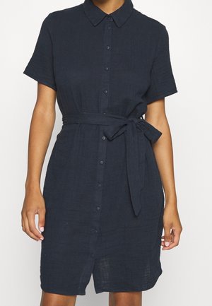 Nainen pukeutuneena tummansiniseen lyhythihaiseen nappikiinniitteiseen mekkoon, jossa on solmittu kangasvyö vyötäröllä, seisoo yksiväristä taustaa vasten.