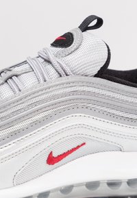 Teniska Nike Air Max 97 v šedé a bílé barvě. Má strukturovaný síťový svršek, charakteristický vlnitý design, červené logo Nike a viditelné vzduchové polstrování.