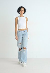 Λευκό ριμπ αμάνικο μπλουζάκι συνδυασμένο με ανοιχτό μπλε χαλαρά τζιν με μεγάλες σχισμές. Το μοντέλο φορά λευκά αθλητικά παπούτσια. Απλή φόντο.