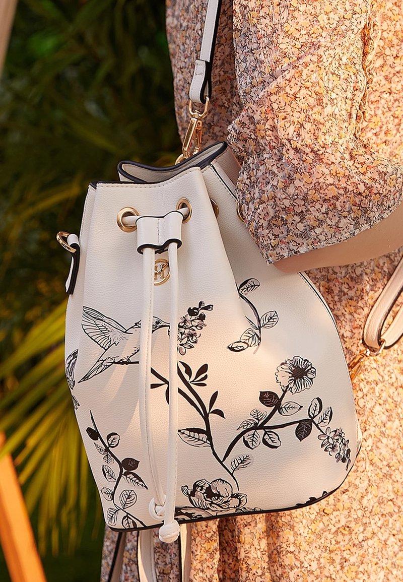 Sac seau blanc avec imprimés floraux et oiseaux noirs porté par une personne en robe fleurie, en extérieur avec des plantes vertes en arrière-plan.