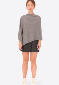 Poncho grigio realizzato in tessuto morbido, caratterizzato da un taglio asimmetrico e un design drappeggiato. Abbinato a una gonna scura e sneaker chiare.