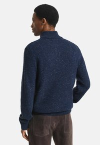 Man staat met de rug naar de camera gekeerd, gekleed in een gestructureerde marineblauwe gebreide trui en donkerbruine broek, tegen een effen lichte achtergrond.