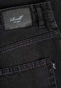 Primer plano del bolsillo trasero de unos vaqueros de mezclilla negros con una etiqueta de marca cosida "Reell est. 1997" en la cinturilla y una pequeña etiqueta de tela en el borde del bolsillo.