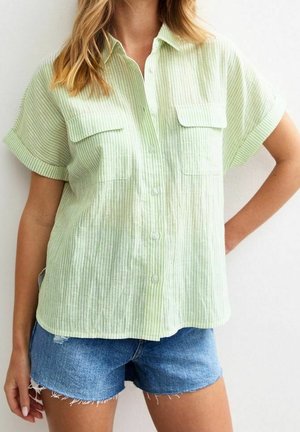 Kobieta ubrana w jasnozieloną koszulę na krótki rękaw z kieszeniami i niebieskie jeansowe szorty, stoi z jedną ręką na biodrze.