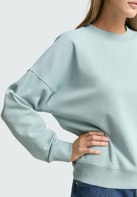 Világoskék pulóver puha anyagból, kerek nyakkivágással, lógó vállakkal és bordás mandzsettákkal, farmernadrággal párosítva.