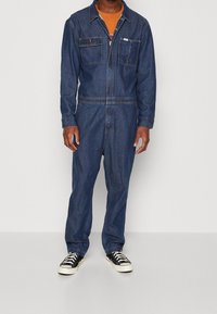 Salopette en jean bleu foncé à manches longues, avec poches poitrine, fermeture éclair et coupe droite, portée sur une chemise orange et des baskets noires.