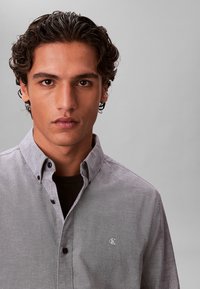 Νέος άνδρας με σγουρά σκουρόχρωμα μαλλιά, φορά γκρι πουκάμισο με κουμπιά πάνω από μαύρο τοπ, κοιτάζει μπροστά σε απλό γκρι φόντο.