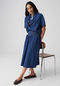 Vrouw leunend op een stoel, gekleed in een blauw denim kort mouwen shirt en bijpassende midi-rok met bruine platte schoenen, voor een effen achtergrond.