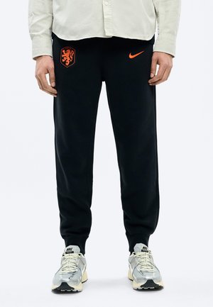 Svarte joggebukser med oransje løvekryss og Nike-logo, båret med hvit skjorte og hvite-grå Nike-sneakers, stående foran en hvit bakgrunn.