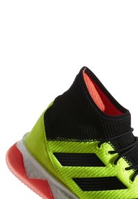 Korkea urheilukenkä, jossa on neonkeltaista ja mustaa neulospintaa, punainen pohjareuna ja mustat nauhat, suunniteltu urheilusuoritusta varten.