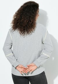 Šedá mikina s dlouhými rukávy, se bílými lemovkami podél ramenních a rukávových švů. Měkká, texturovaná látka, uvolněný střih.