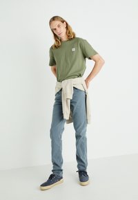 T-shirt in cotone verde oliva con piccolo logo, maglione beige chiaro legato in vita, pantaloni in denim blu e sneakers in suede blu.