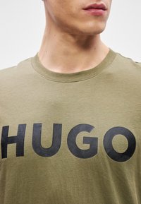 Olive zelené bavlnené tričko s veľkým čiernym logom "HUGO" na prednej strane. Okrúhly výstrih a krátke rukávy s hladkou textúrou.
