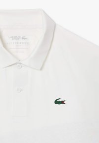 Hvid polo-shirt med en tætsiddende design, tre-knap stolpe og et grønt broderet krokodillelogo på venstre bryst. Glat stoftekstur.