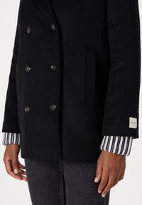 Zwarte wollen mantel met dubbele knoopsluiting, zijzak en gestreepte gebreide manchetten. Calvin Klein-label op de manchette.