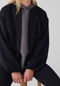 Personne assise, les mains jointes, portant un sweat à capuche gris sous une veste noire ample et un pantalon noir sur un fond uni.