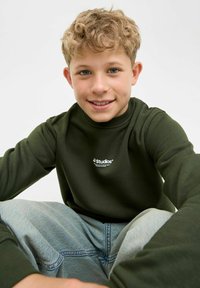 Zielony sweatshirt z długim rękawem i okrągłym dekoltem, z białym logo na piersi, zestawiony z jasnoniebieskimi dżinsami. Miękka, teksturowana tkanina.