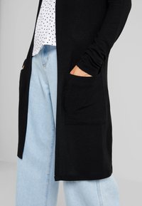 Black long cardigan with pockets, worn over a white polka-dot top and light blue wide-leg jeans. Soft fabric, open front design.