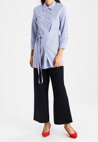 Pasiasta niebieska długa koszula z wiązaniem w talii, rękawy do trzech ćwiartek, w połączeniu z szerokimi czarnymi spodniami i czerwonymi balerinami. Prosty krój kołnierza.