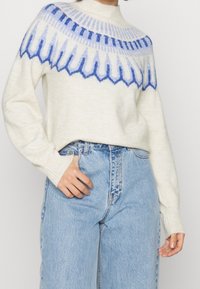 Woman wearing a cream sweater with blue geometric patterns and high-waisted light blue jeans, hand resting on pocket.
