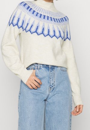 Femme portant un pull crème avec des motifs géométriques bleus et un jean taille haute bleu clair, la main posée sur la poche.