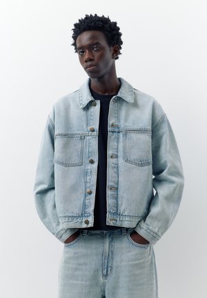 Młody mężczyzna z rękami w kieszeniach, ubrany w jasnoniebieską kurtkę dżinsową i dopasowane dżinsy, na czarnej koszulce, stojący na białym tle.