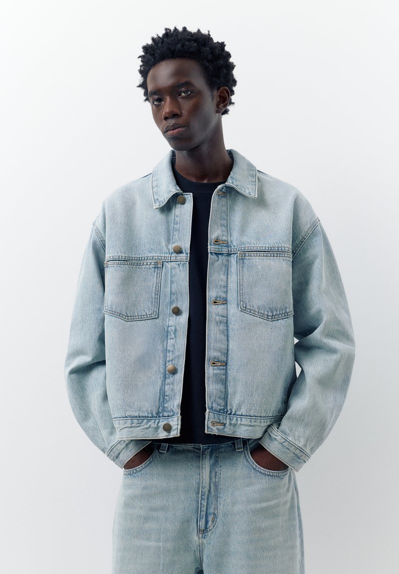Jeune homme les mains dans les poches, portant une veste en jean bleu clair et un jean assorti sur un t-shirt noir, debout devant un fond blanc.