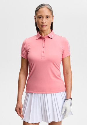 Vrouw die een roze poloshirt met korte mouwen, een witte geplooide rok en een witte golfhandschoen aan haar linkerhand draagt, staand tegen een effen achtergrond.