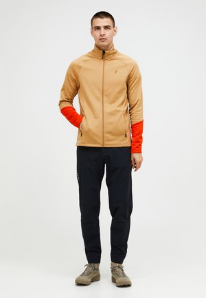 Beige Jacke mit orangen Ärmeln, Reißverschluss und Logodetail. Getragen mit dunklen Hosen und hellbraunen Schuhen. Glattes Material, sportliche Passform.