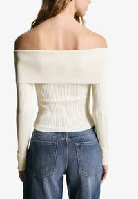 Cream off-shoulder ribbed sweater with long sleeves, fitted body, and textured knit; paired with high-waisted blue denim jeans.