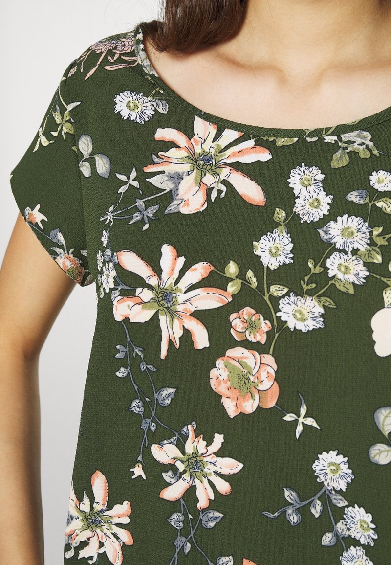 Femme portant un haut vert à manches courtes avec un motif floral en blanc, pêche et vert clair.