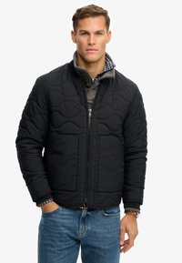 Czarna pikowana kurtka na zamek błyskawiczny, z prążkowanym kołnierzem i dwoma przednimi kieszeniami. Noszona na szarej koszuli w kratę.