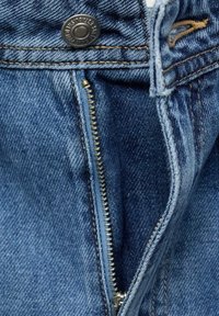 Detalj av denimjeans med dragkedja, som har en metalldragkedja med mässingskäftar, mörkblåt tyg och en knappstängning i midjan.